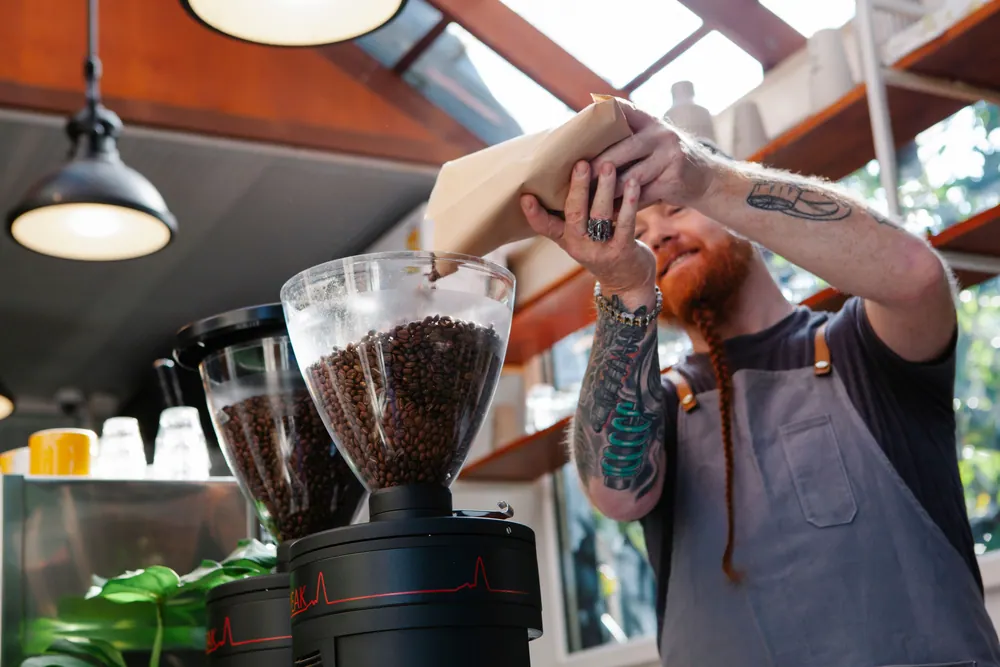 Tattooed barista replenishing coffee grinder beans in a modern café setting.