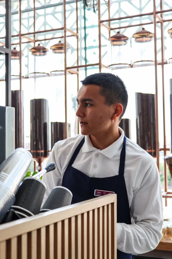 Young male barista working in a trendy café setting, wearing an apron.