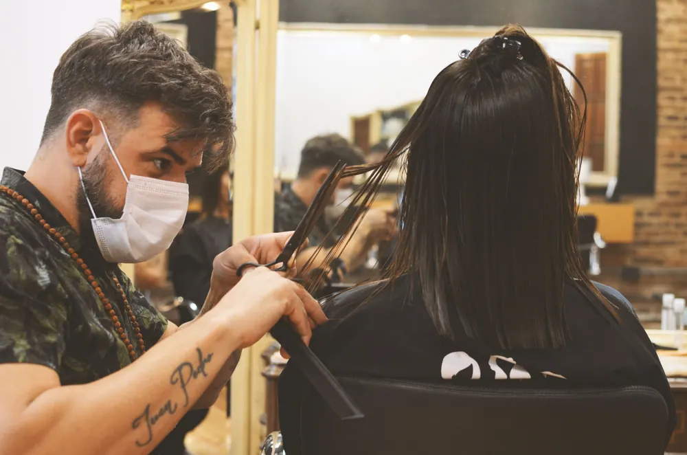 Barber wearing facemask carefully cuts woman's hair indoors, ensuring safety and style in a barbershop.