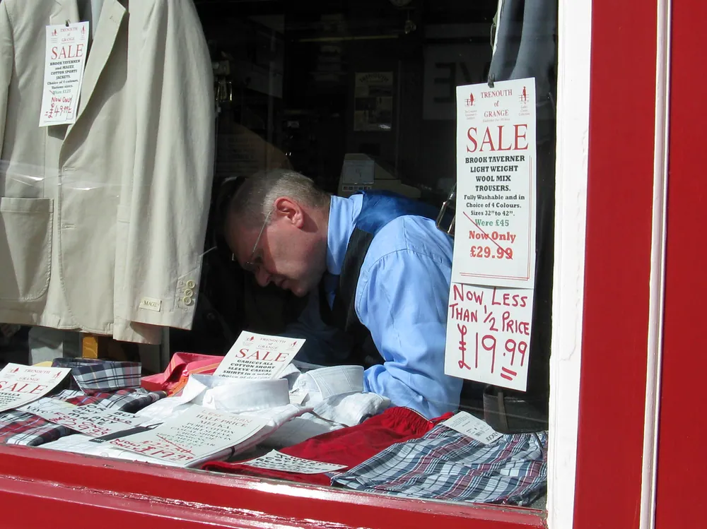 A man in a shop window arranging clothes for a sale with various sale signs displayed.
