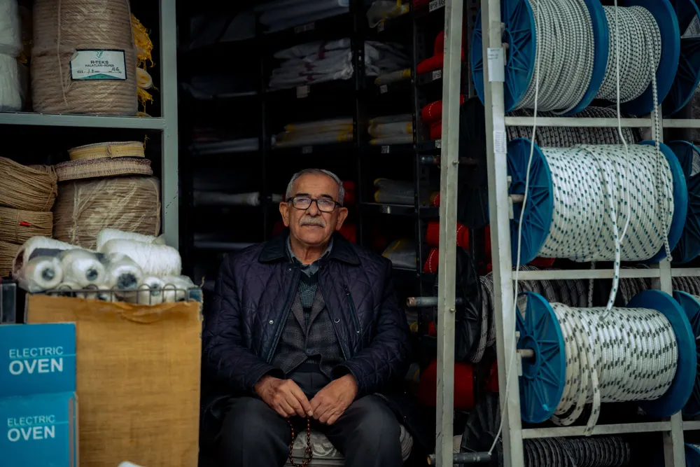 An elderly man sitting inside a shop surrounded by various coiled ropes and textile materials.