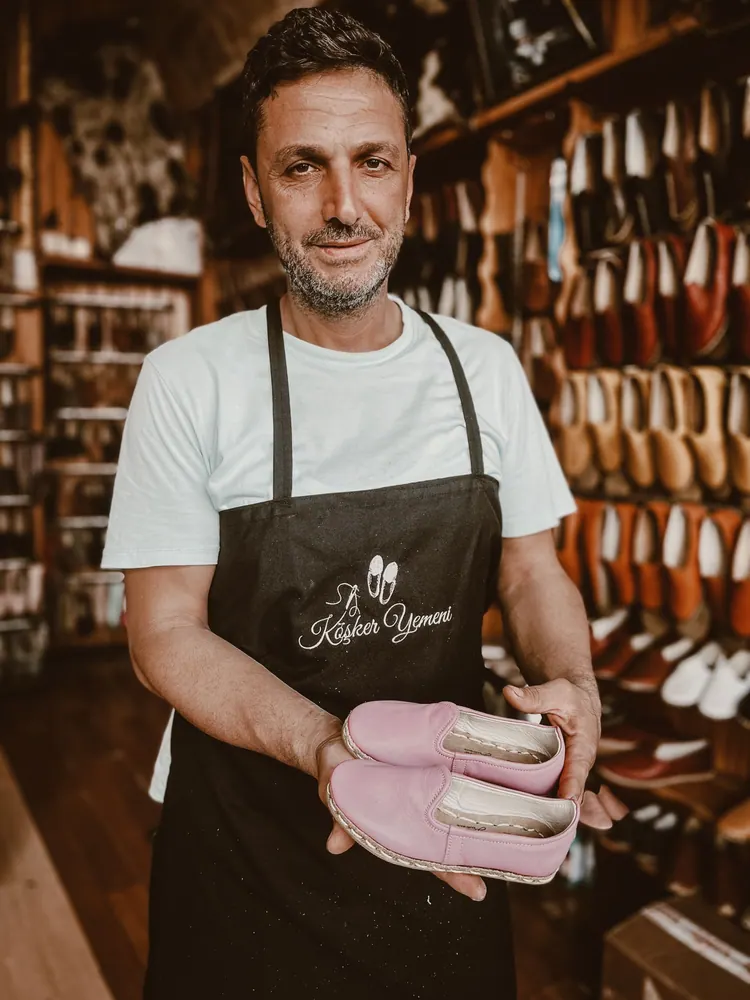 A smiling craftsman in a shoe store holding pink handmade leather shoes, showcasing his craftsmanship.