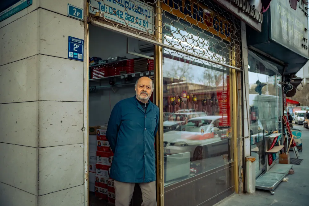 A senior male shopkeeper standing at his store entrance on a busy city street, looking outwards.