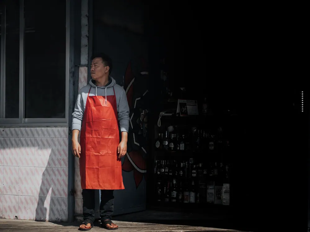 A man wearing a red apron stands outside a shop on a city street, partially in shadow.