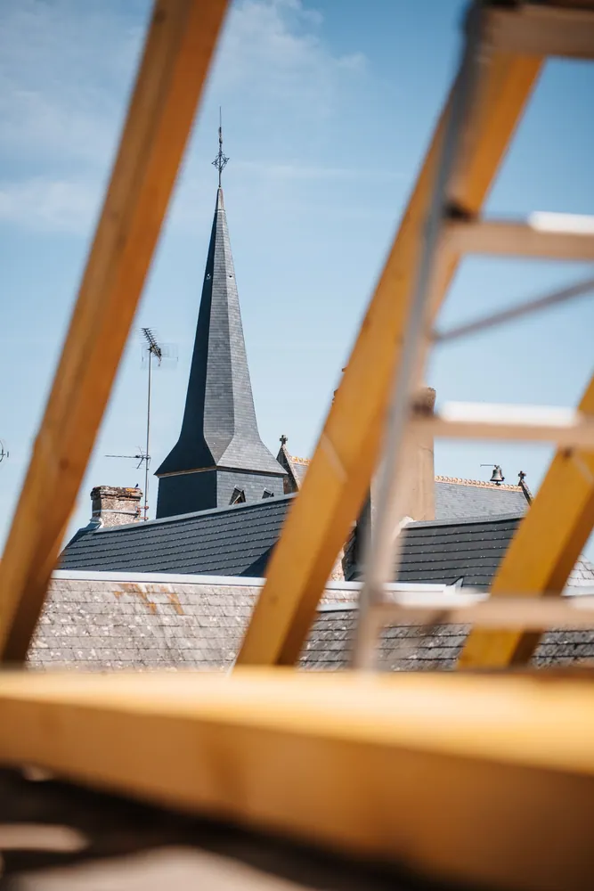 View of a church spire through the framing of a rooftop construction site under clear skies.
