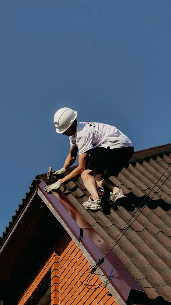 A roofer in a safety helmet renovating a brick house roof under clear blue skies.