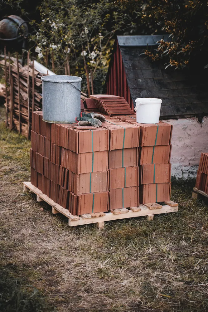 Pallet of stacked roof tiles with metal and plastic buckets outside near a shed.