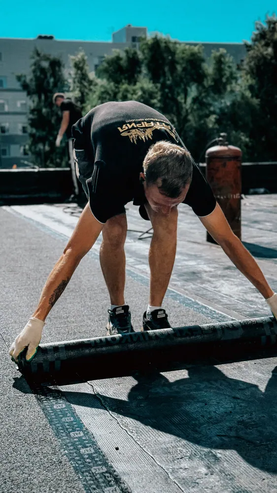 A construction worker applies waterproof membrane on a flat roof under bright daylight.