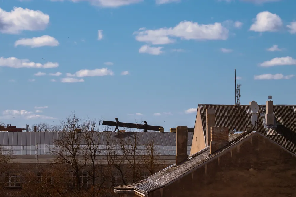 Silhouette of construction workers on a roof under a bright blue sky with clouds.