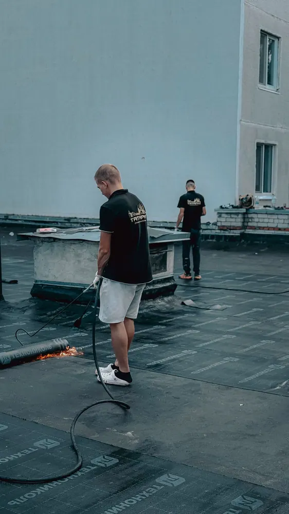 Two contractors wear safety equipment and install roofing on a flat building roof with a torch.