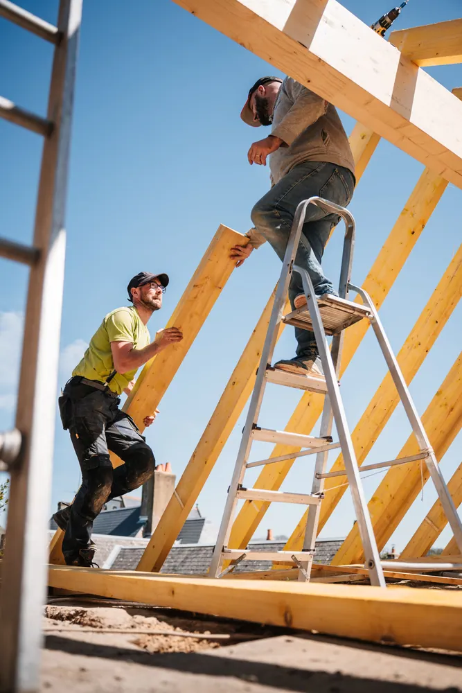 Two construction workers building a wooden roof structure on a sunny day.