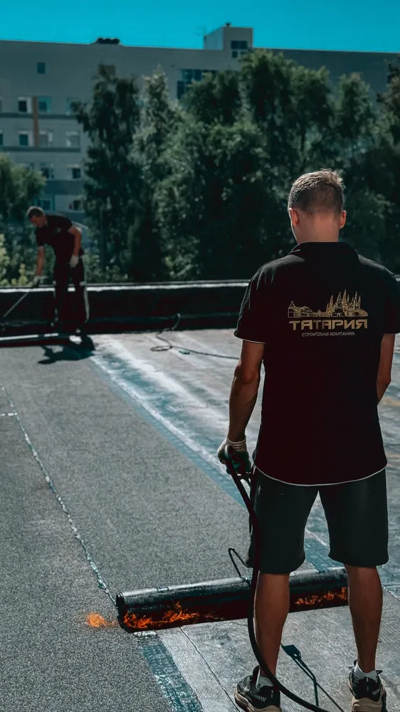 Two men applying hot tar on a flat roof under a bright sunny day, showcasing roofing expertise.