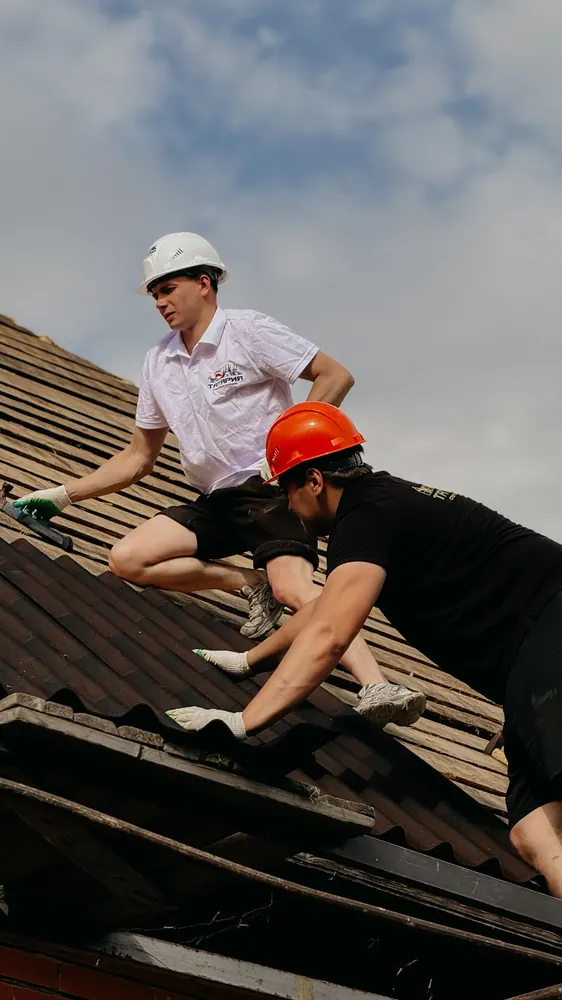 Two construction workers in safety helmets repairing a roof under clear skies.