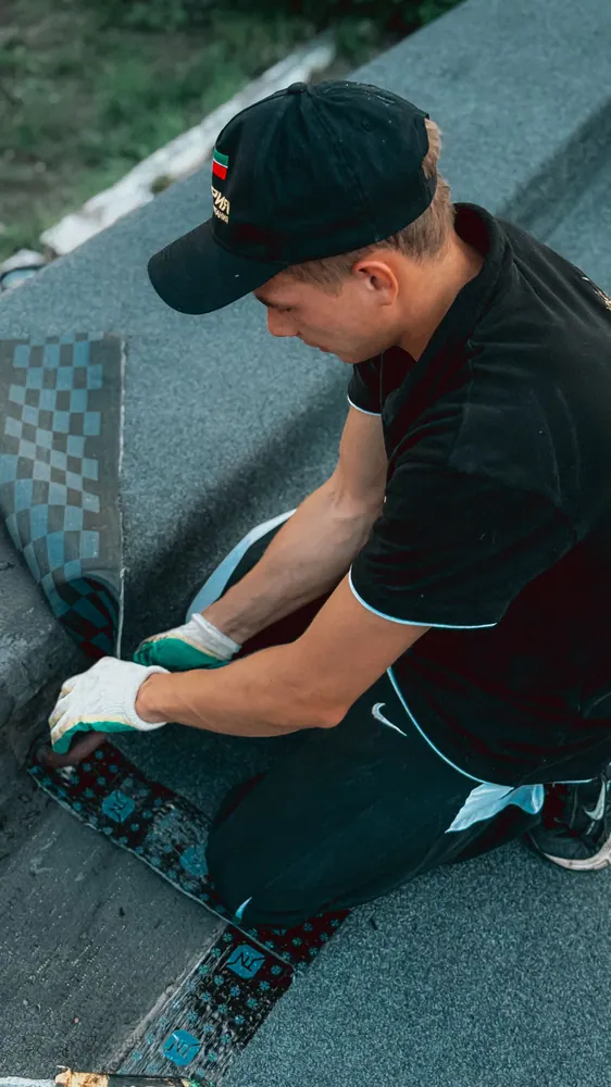 Man wearing gloves and a cap installing shingles on a flat roof on a sunny day.